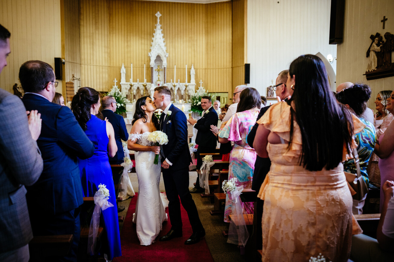 Tara and Darren’s Unforgettable Wedding at Bellingham Castle 84 A bride and groom kiss at the altar of a church during their beautiful wedding at Bellingham Castle, as guests stand and watch, with elegant wedding decorations and flowers in the background.
