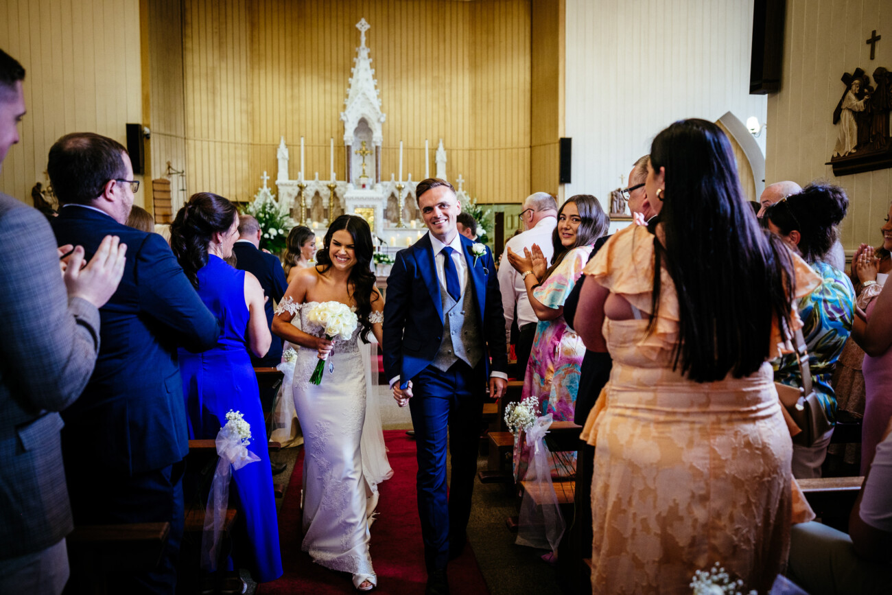 Tara and Darren’s Unforgettable Wedding at Bellingham Castle 85 A bride and groom walk down the aisle together in a church, smiling and holding hands, as guests stand and applaud, capturing the joy of their beautiful wedding at Bellingham Castle.