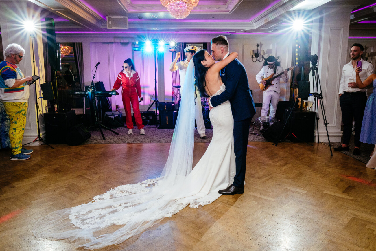 Tara and Darren’s Unforgettable Wedding at Bellingham Castle 157 Two brides in wedding dresses share a kiss on the dance floor during their wedding at Bellingham Castle, surrounded by musicians and guests in a brightly lit, elegant room.