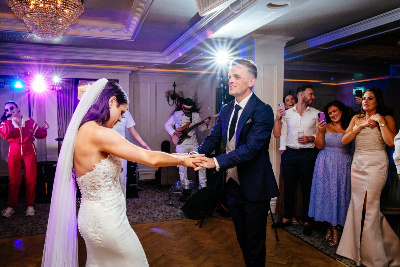 Tara and Darren’s Unforgettable Wedding at Bellingham Castle 158 A bride and groom dance together at their wedding reception at Bellingham Castle while guests watch and a live band performs in the background.