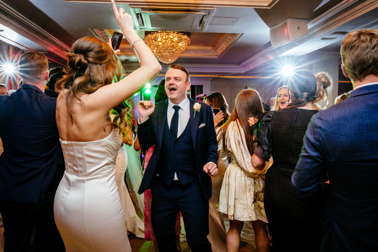Tara and Darren’s Unforgettable Wedding at Bellingham Castle 159 A group of people in formal attire dance and celebrate at an indoor event, with colourful lights and a chandelier overhead, capturing the joyous atmosphere of a wedding at Bellingham Castle.