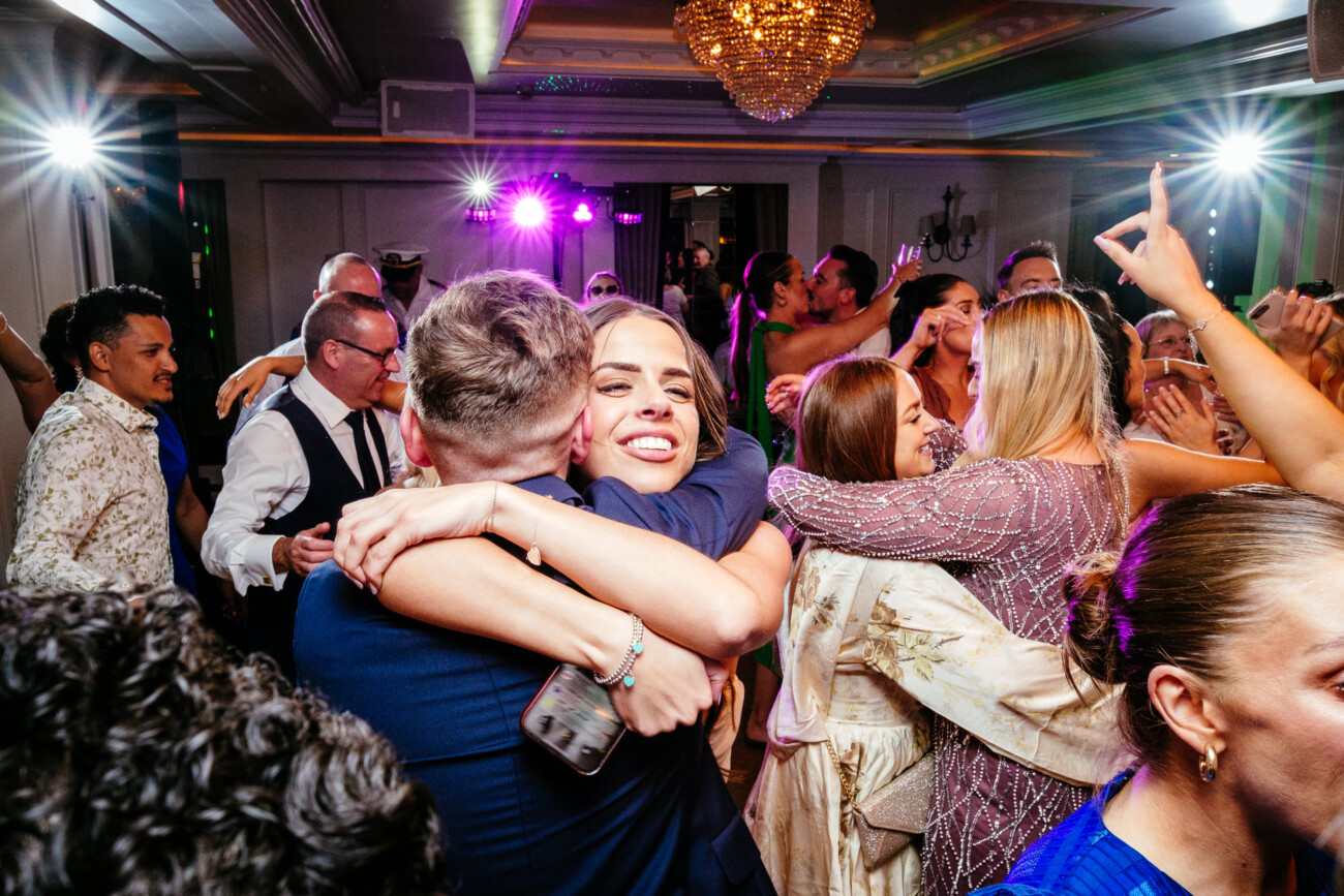 Tara and Darren’s Unforgettable Wedding at Bellingham Castle 160 A woman smiles as she hugs a man in a crowded room with people dancing and celebrating under bright lights during a joyful wedding at Bellingham Castle.