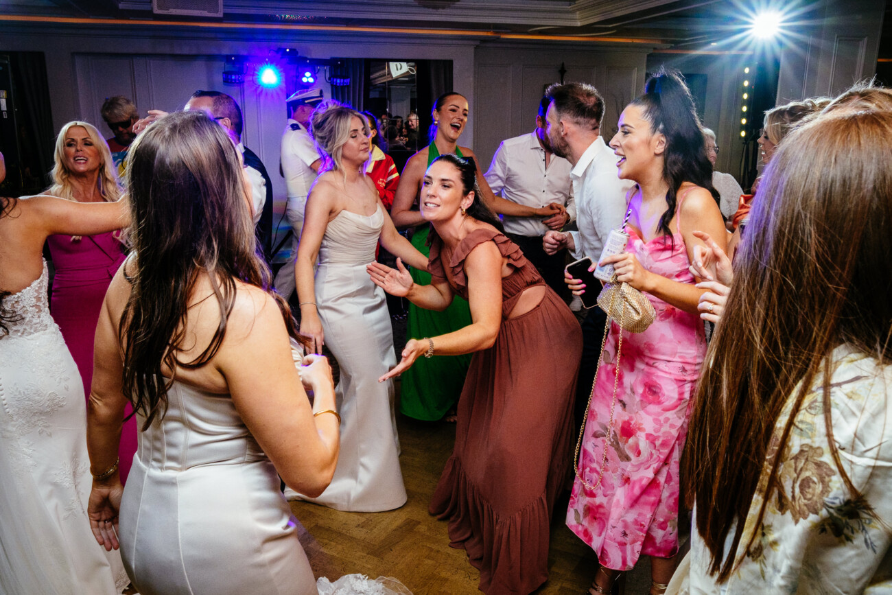 Tara and Darren’s Unforgettable Wedding at Bellingham Castle 161 A group of people in formal attire dance and laugh together at an indoor party, with colourful lights illuminating the scene, celebrating a joyous wedding at Bellingham Castle.