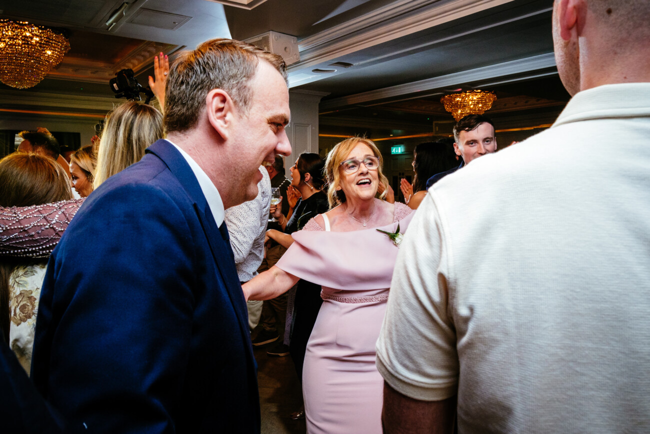 Tara and Darren’s Unforgettable Wedding at Bellingham Castle 162 A group of people, including a woman in a light pink dress and a man in a navy suit, socialising and smiling at an elegant wedding at Bellingham Castle, with chandeliers overhead creating a warm and inviting atmosphere.