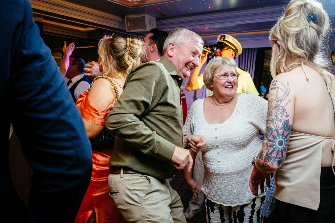 Tara and Darren’s Unforgettable Wedding at Bellingham Castle 163 A group of adults dance and socialise at an indoor party during a wedding at Bellingham Castle, smiling and interacting under colourful lights.