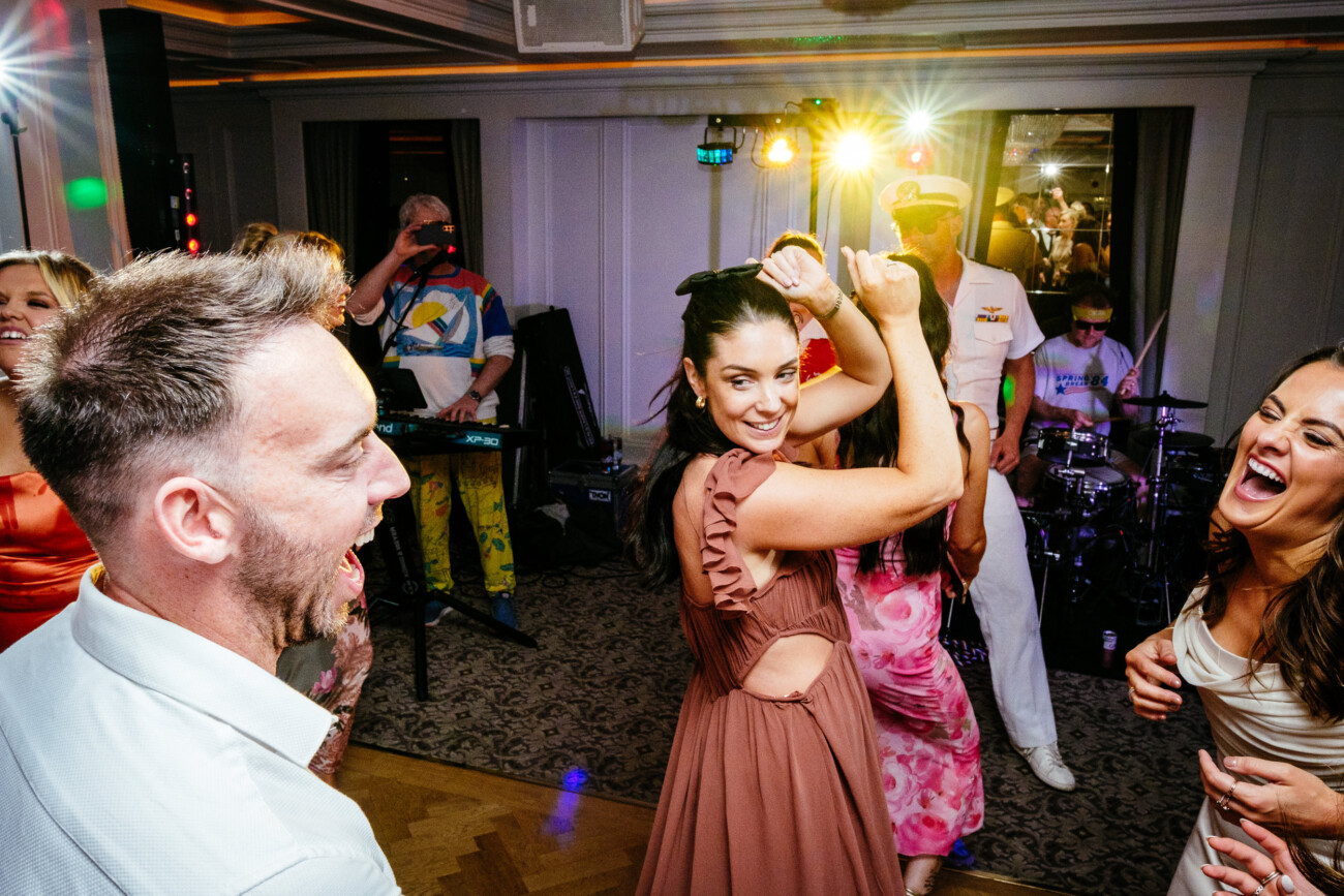 Tara and Darren’s Unforgettable Wedding at Bellingham Castle 164 People dancing and laughing in a brightly lit indoor setting, with a live band playing in the background during a wedding at Bellingham Castle.