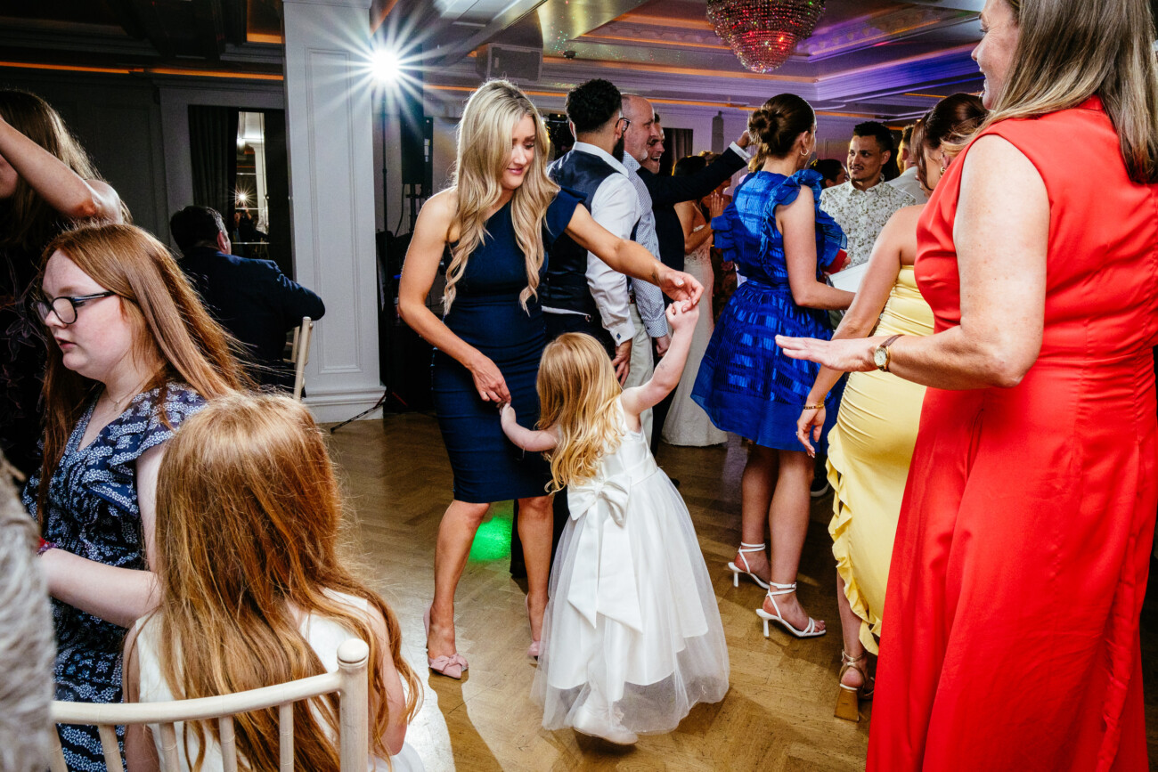 Tara and Darren’s Unforgettable Wedding at Bellingham Castle 165 At a wedding at Bellingham Castle, a woman in a blue dress dances with a young girl in a white dress at a lively indoor party with other guests and colourful lighting.