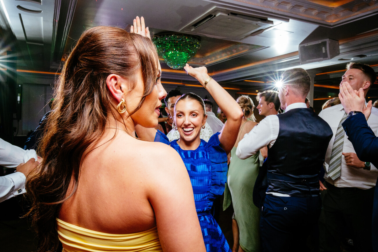 Tara and Darren’s Unforgettable Wedding at Bellingham Castle 166 People in formal attire dancing and celebrating at an indoor event, with a woman in a blue dress raising her arms and smiling at another woman in a yellow dress—capturing the joyful spirit of a wedding at Bellingham Castle.