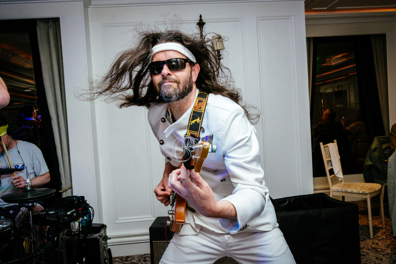 Tara and Darren’s Unforgettable Wedding at Bellingham Castle 167 Man with long hair, sunglasses, and a headband plays electric guitar energetically in a white suit indoors during a lively wedding at Bellingham Castle, with people and musical equipment in the background.