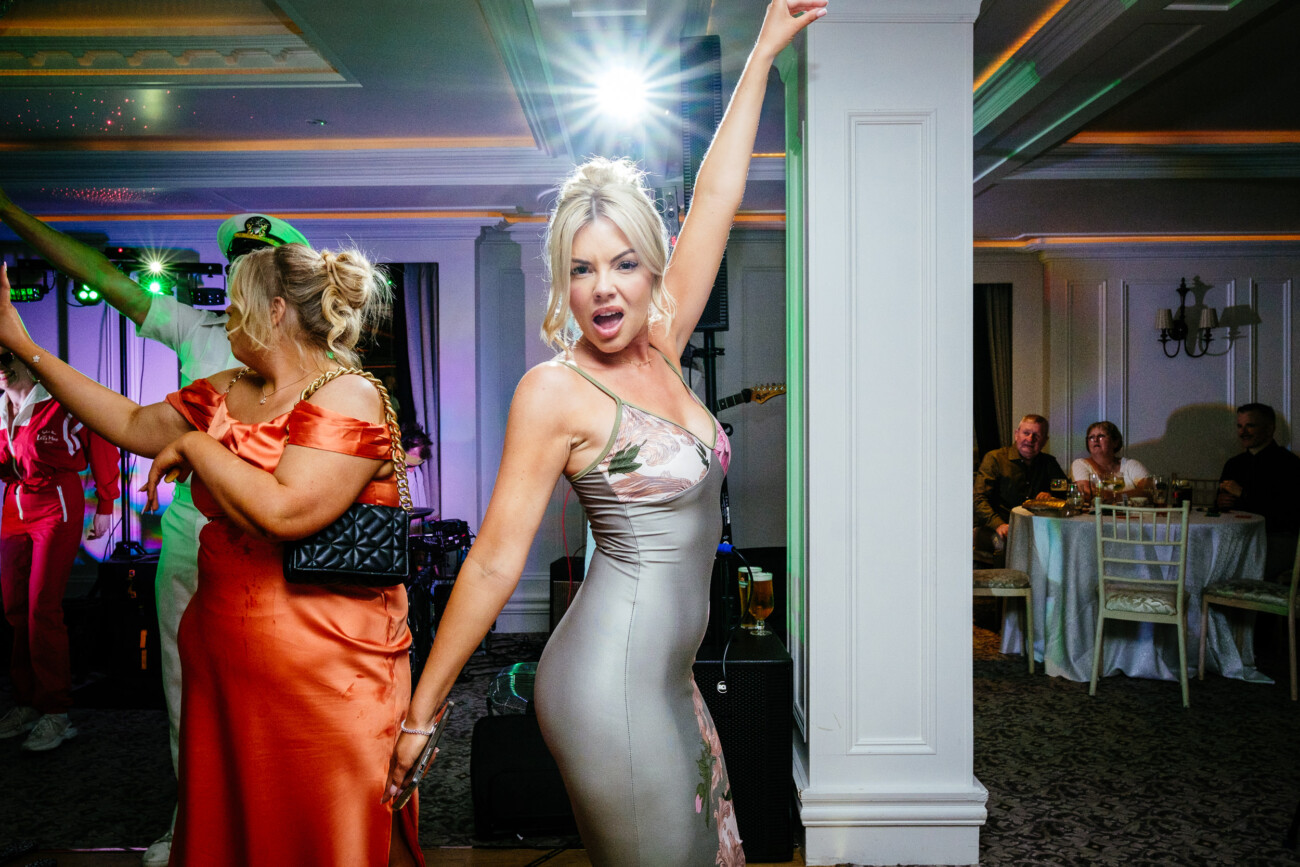 Tara and Darren’s Unforgettable Wedding at Bellingham Castle 168 A woman in a silver dress poses with one arm raised at a lively indoor event during a wedding at Bellingham Castle. Other guests are visible in the background, some seated at tables.