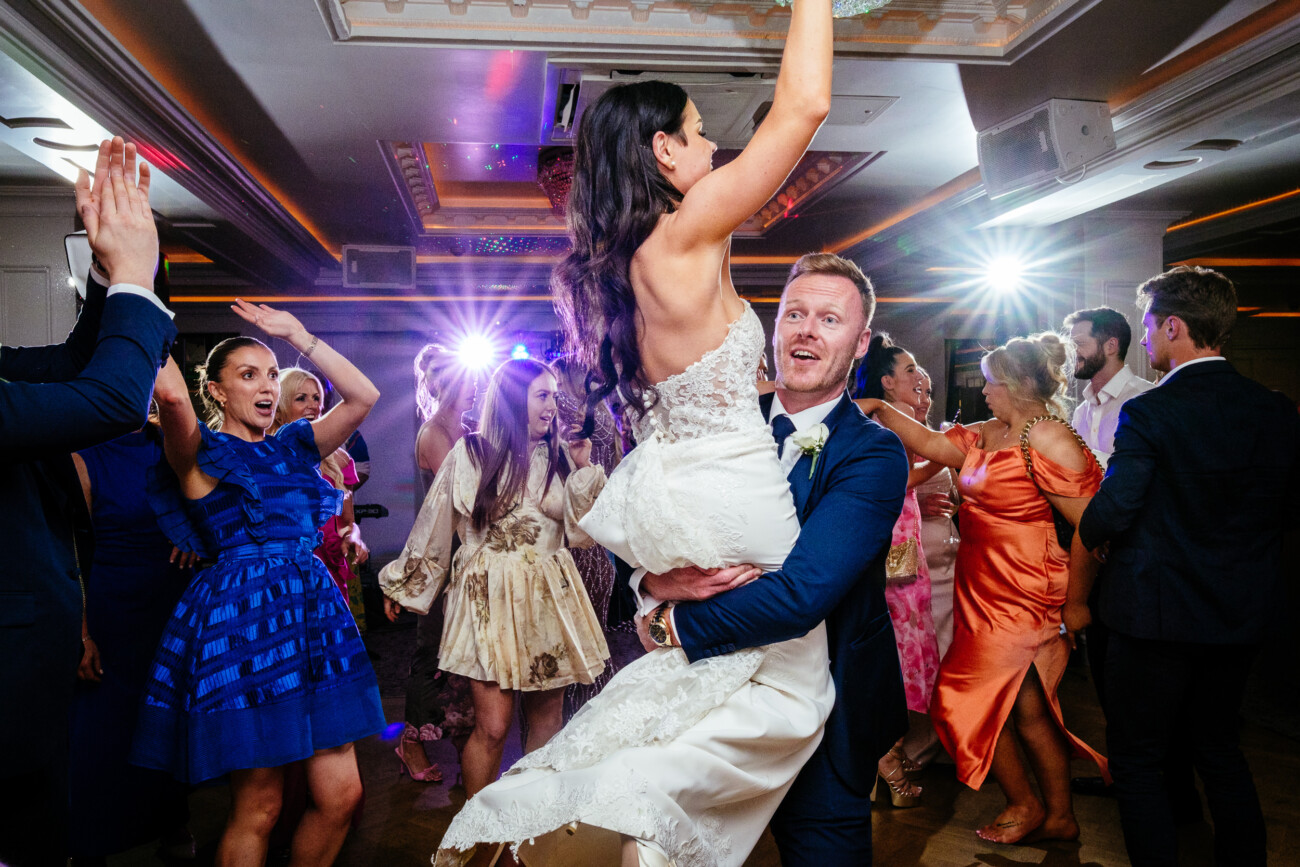 Tara and Darren’s Unforgettable Wedding at Bellingham Castle 169 A groom lifts a bride as they dance at their wedding at Bellingham Castle, surrounded by wedding guests in colourful attire on a lively dance floor with bright lights.