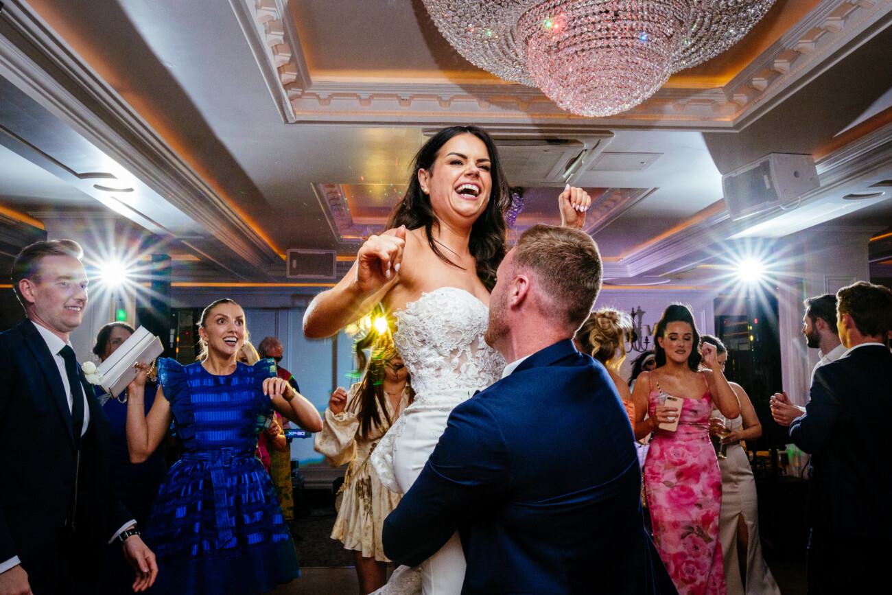 Tara and Darren’s Unforgettable Wedding at Bellingham Castle 170 A groom lifts the bride in celebration on the dance floor during a wedding at Bellingham Castle, surrounded by cheering guests in formal attire beneath a large chandelier.