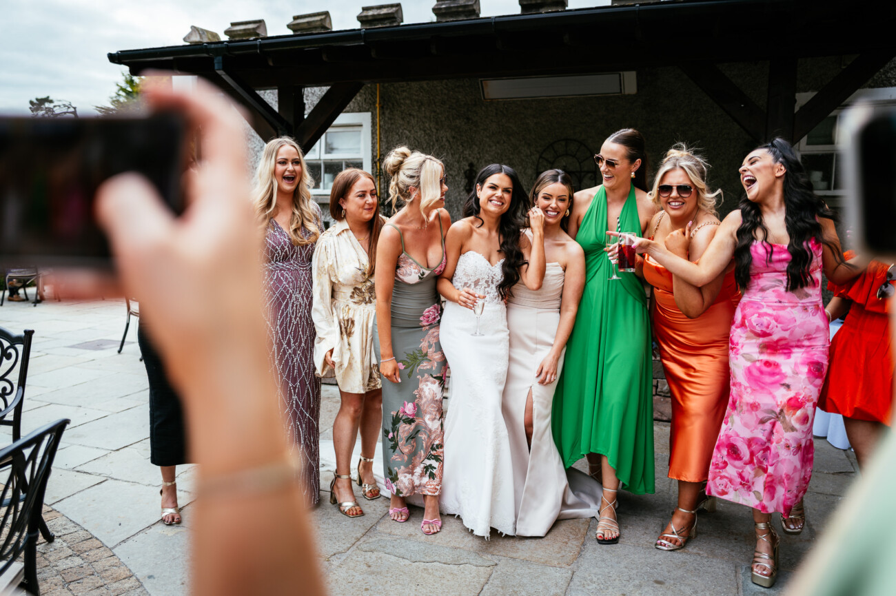 Tara and Darren’s Unforgettable Wedding at Bellingham Castle 115 A bride in a white dress poses and smiles with seven women in colourful dresses while someone takes a photo outside at an event during a beautiful wedding at Bellingham Castle.