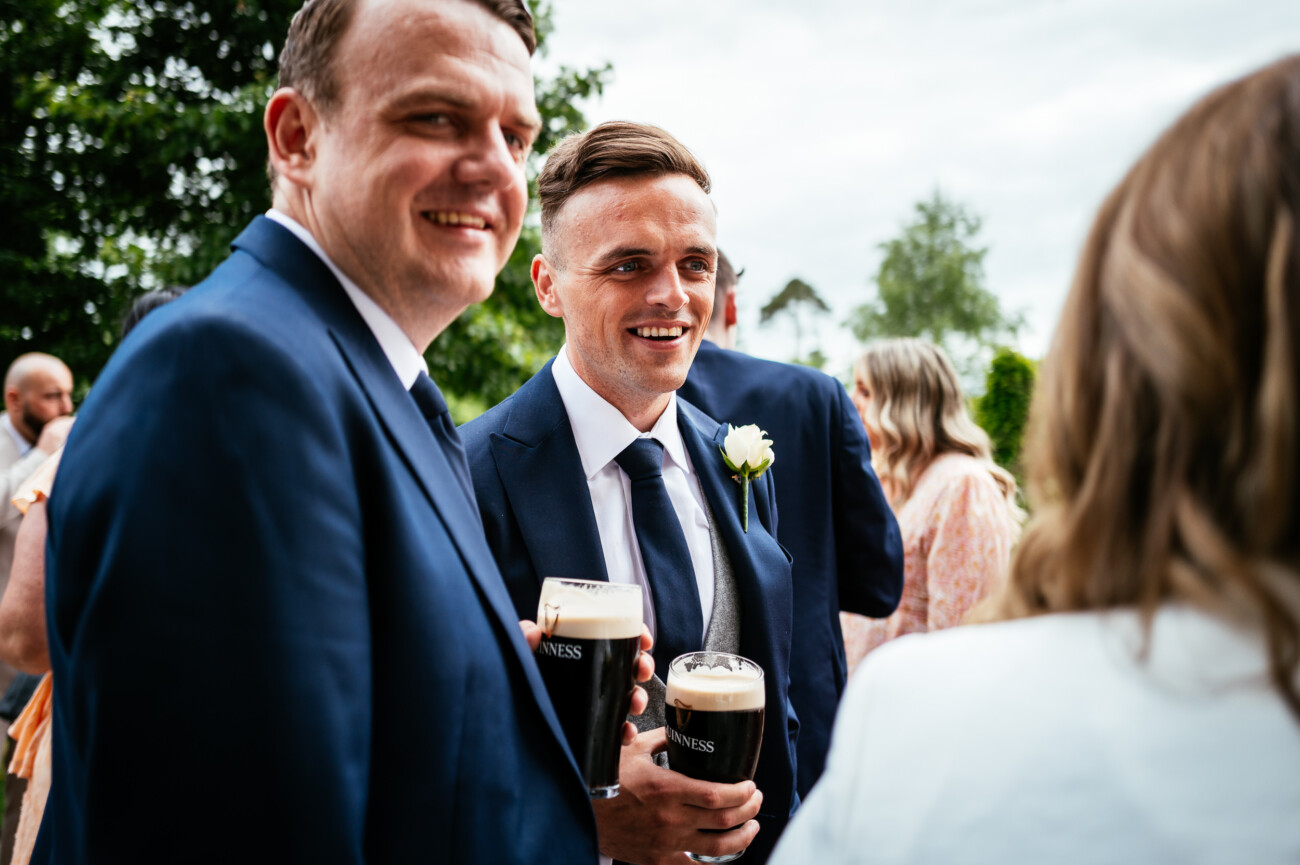 Tara and Darren’s Unforgettable Wedding at Bellingham Castle 116 Two men in suits holding pints of Guinness stand outdoors, smiling and talking with a woman during a wedding at Bellingham Castle. Other people and trees are visible in the background.