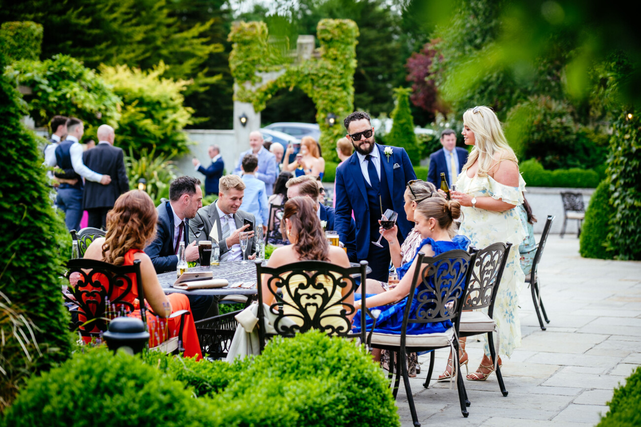 Tara and Darren’s Unforgettable Wedding at Bellingham Castle 117 A group of people in formal attire sit and stand around outdoor tables in a lush garden setting, talking and enjoying drinks during a beautiful wedding at Bellingham Castle.