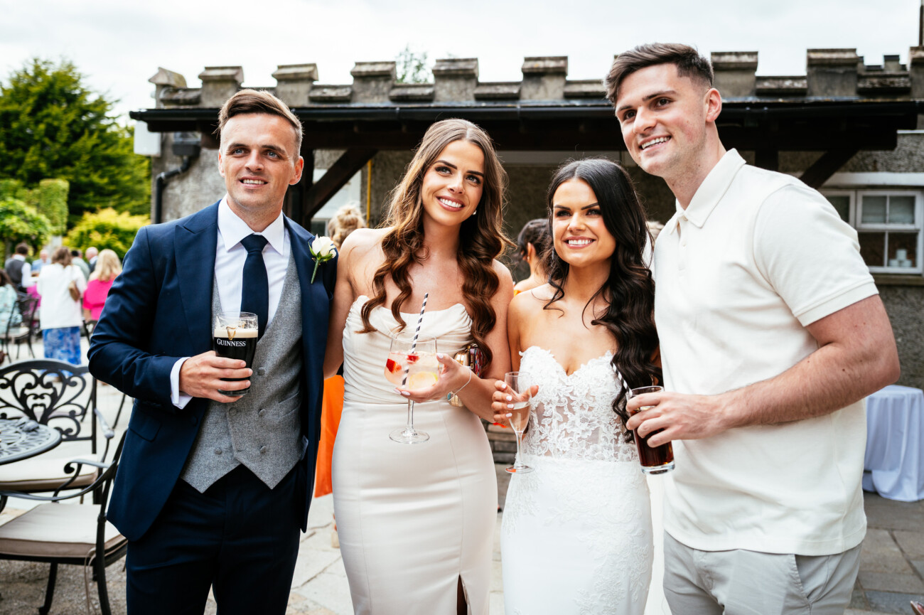 Tara and Darren’s Unforgettable Wedding at Bellingham Castle 119 Four people posing together outdoors at a formal event, likely a wedding at Bellingham Castle, with two women in white dresses and two men—one in a suit and the other in a white shirt—all holding drinks and smiling at the camera.