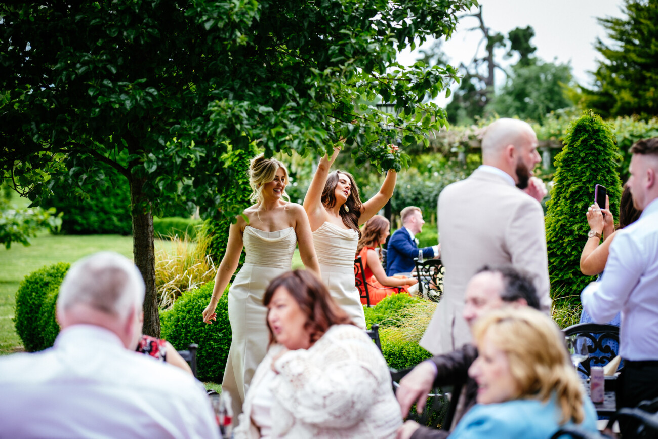 Tara and Darren’s Unforgettable Wedding at Bellingham Castle 120 Two women in formal dresses walk through a garden whilst guests sit and stand around them at an outdoor wedding at Bellingham Castle.
