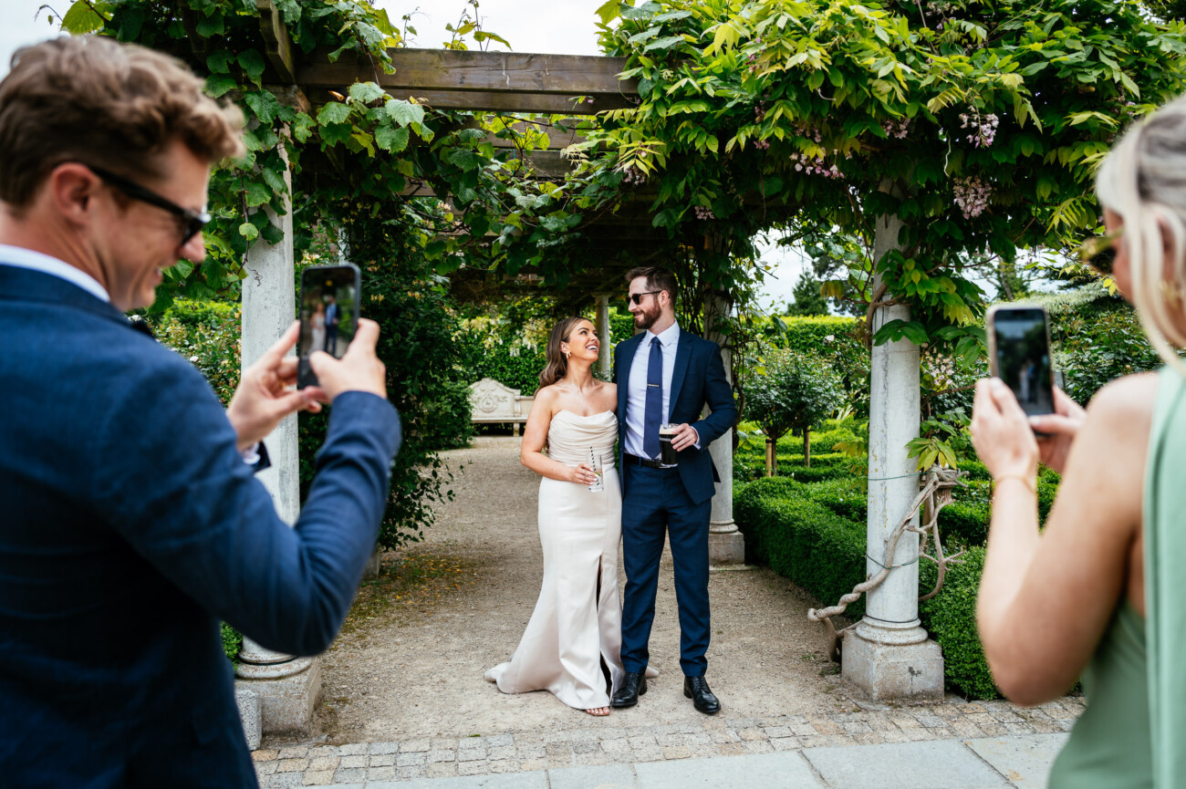 Tara and Darren’s Unforgettable Wedding at Bellingham Castle 121 A bride and groom stand under a vine-covered pergola at their wedding at Bellingham Castle, smiling at each other, whilst guests on either side take photos with their mobiles.