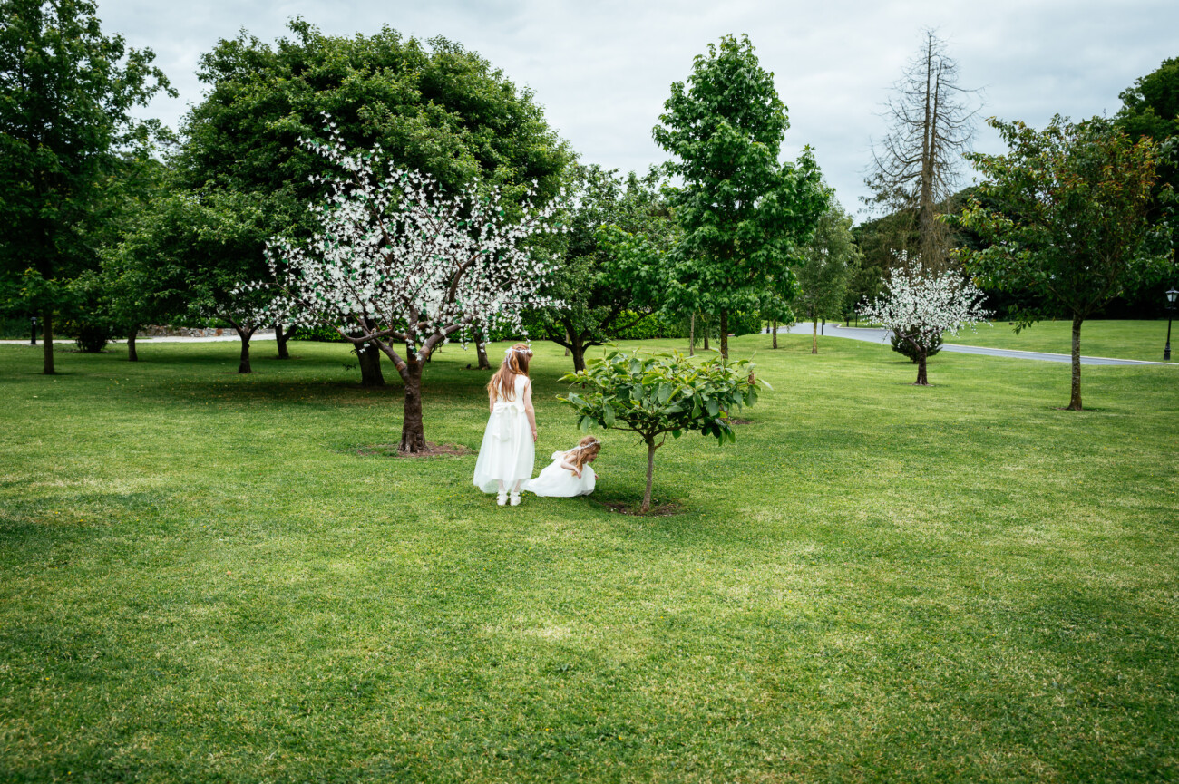 Tara and Darren’s Unforgettable Wedding at Bellingham Castle 122 Two girls in white dresses are in a grassy park with blossoming and leafy trees; one girl stands while the other sits under a small tree, evoking the dreamy atmosphere of a wedding at Bellingham Castle.