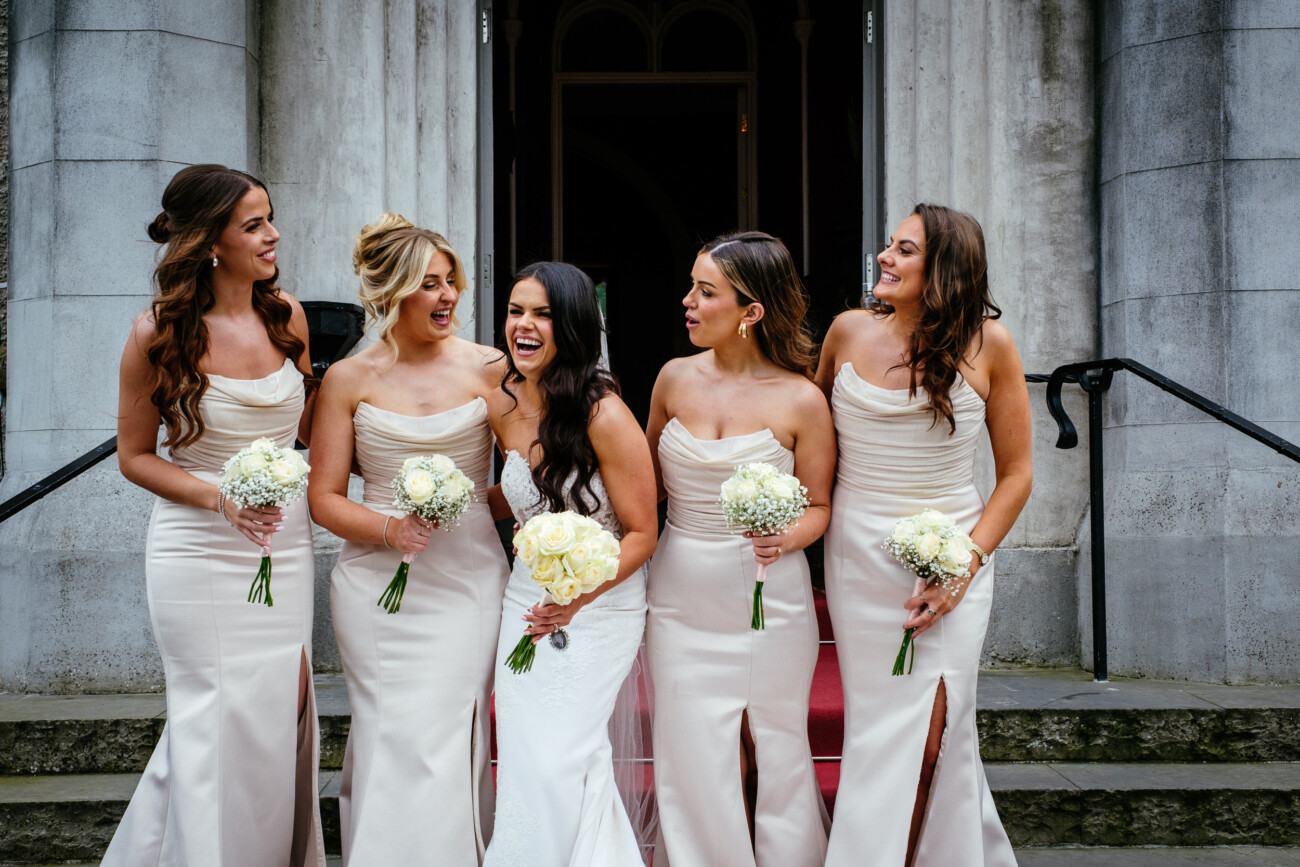 Tara and Darren’s Unforgettable Wedding at Bellingham Castle 123 Five women in matching light-coloured dresses, holding bouquets, stand together smiling on stone steps in front of a building entrance, capturing a joyful moment from a wedding at Bellingham Castle.