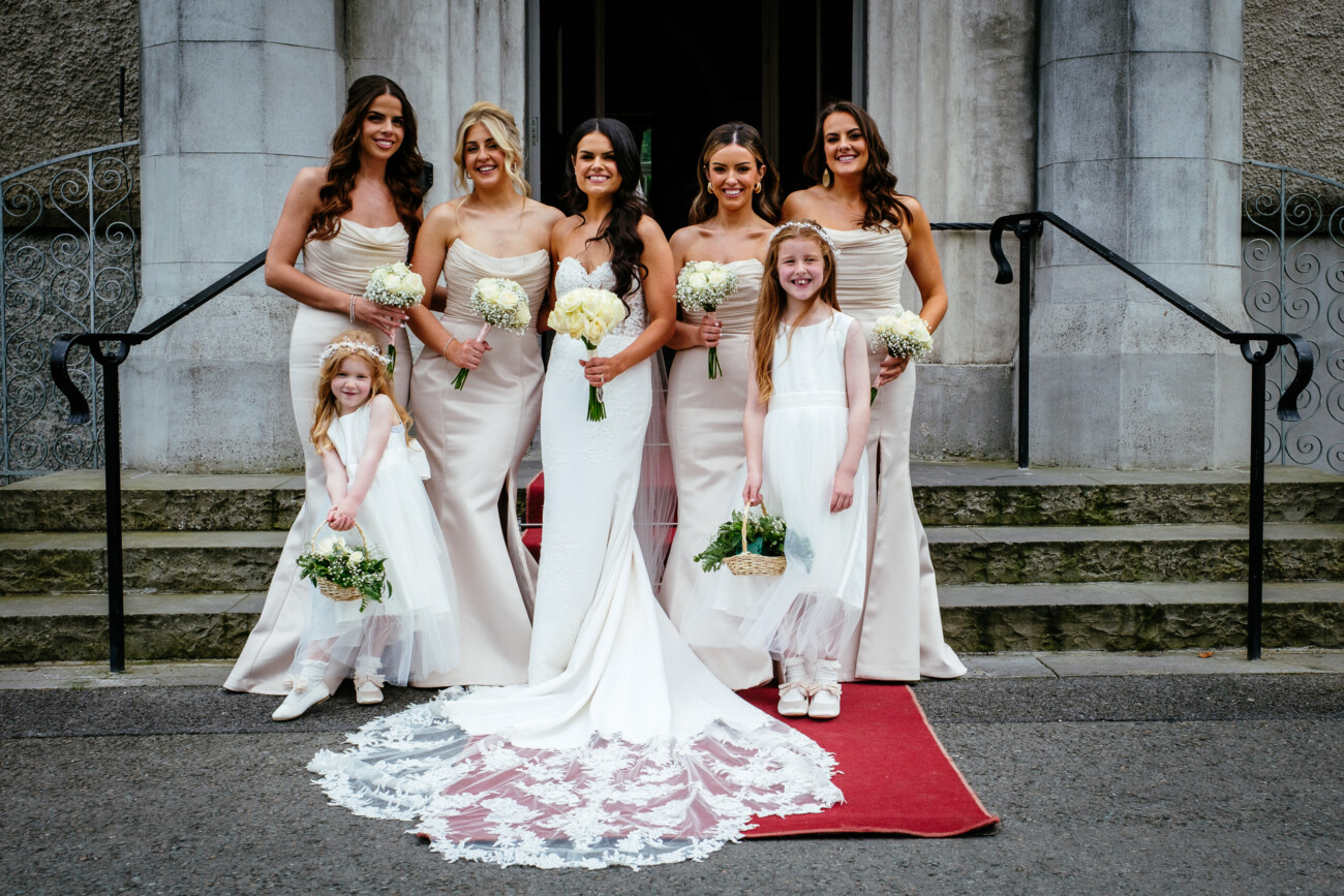 Tara and Darren’s Unforgettable Wedding at Bellingham Castle 124 Six women and girls in formal dresses stand on steps outside a building, with the bride in the centre holding a bouquet, flanked by bridesmaids and flower girls on a red carpet, capturing the elegance of a wedding at Bellingham Castle.