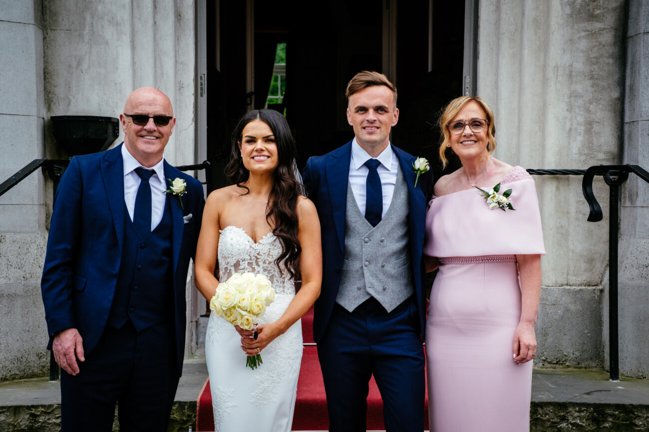 Tara and Darren’s Unforgettable Wedding at Bellingham Castle 125 A bride and groom stand with two adults, all dressed formally, posing for a photo on steps with a red carpet in front of a stone building during their wedding at Bellingham Castle.
