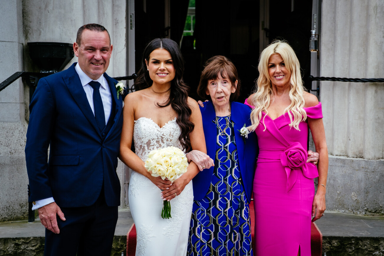 Tara and Darren’s Unforgettable Wedding at Bellingham Castle 126 Four people posing for a formal photo at a wedding at Bellingham Castle; the bride holds a bouquet and stands between an older woman and two adults in formal attire.