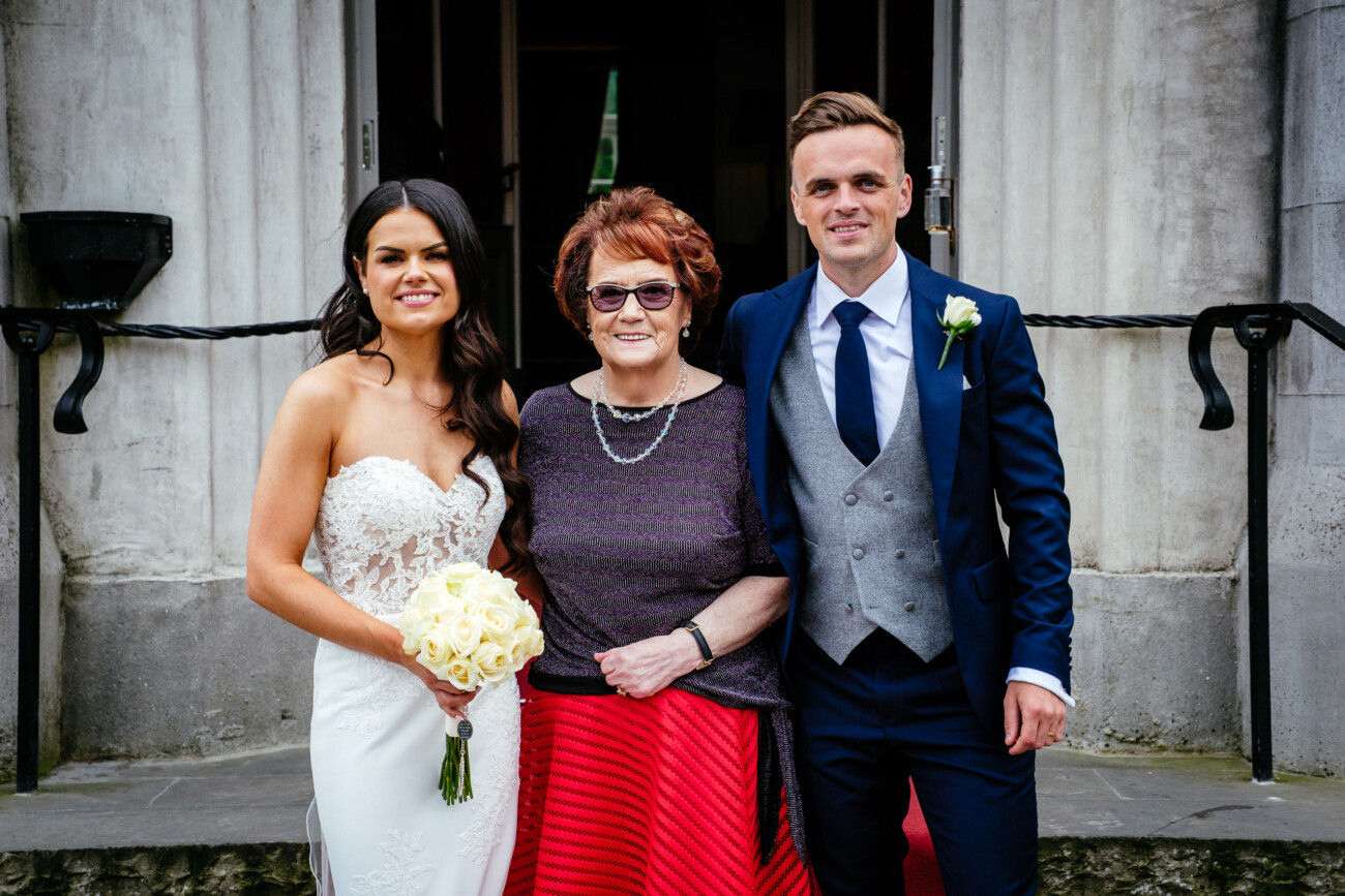 Tara and Darren’s Unforgettable Wedding at Bellingham Castle 127 A bride holding a bouquet, an older woman, and a man in formal attire stand together outside a building, posing for a photo during a wedding at Bellingham Castle.