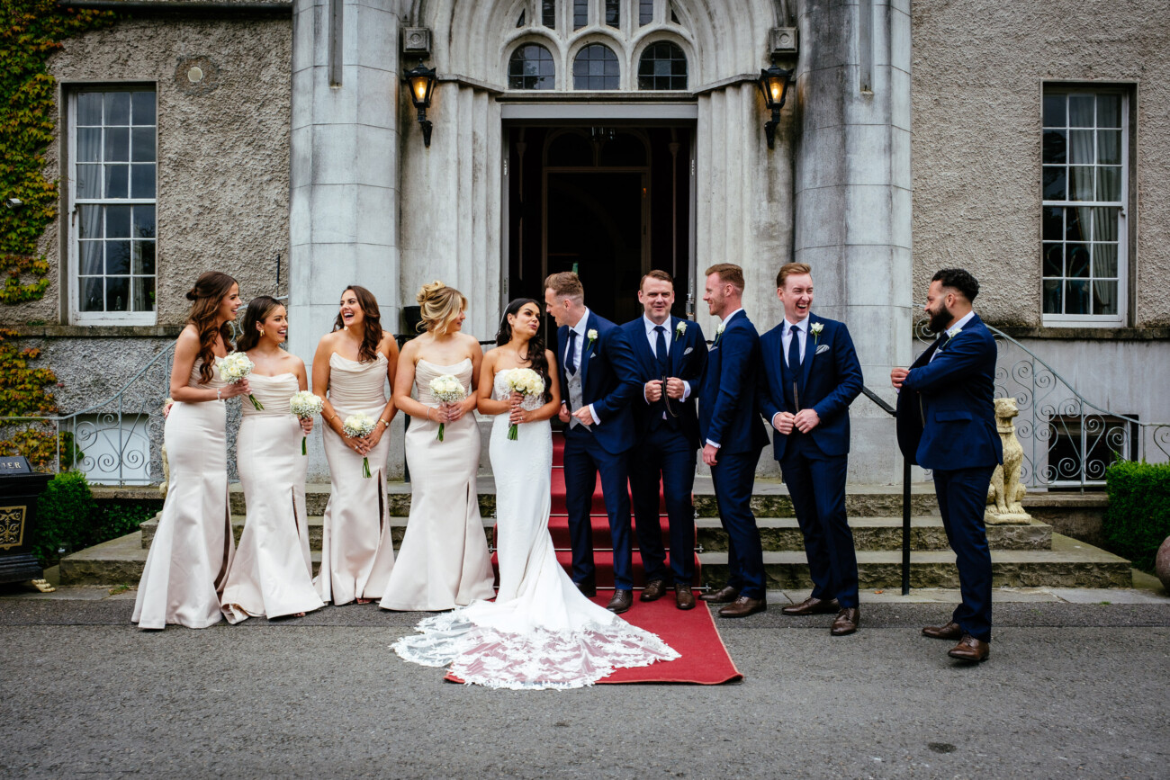 Tara and Darren’s Unforgettable Wedding at Bellingham Castle 128 The bride, groom, bridesmaids, and groomsmen laugh together on the steps of Bellingham Castle after the ceremony.