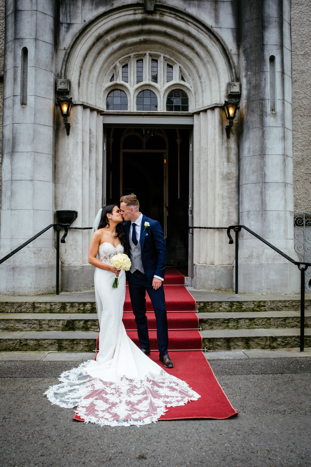 Tara and Darren’s Unforgettable Wedding at Bellingham Castle 129 A bride and groom kiss on the steps of a stone building with arched doors, standing on a red carpet during their wedding at Bellingham Castle.