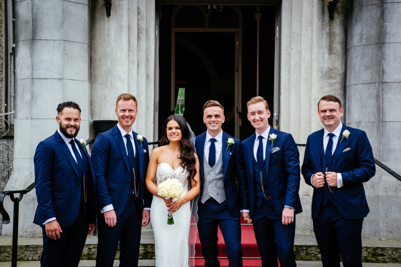 Tara and Darren’s Unforgettable Wedding at Bellingham Castle 130 Bride stands with five men in suits in front of a building entrance with red carpeted steps, all smiling and posing for a wedding photo at Bellingham Castle.
