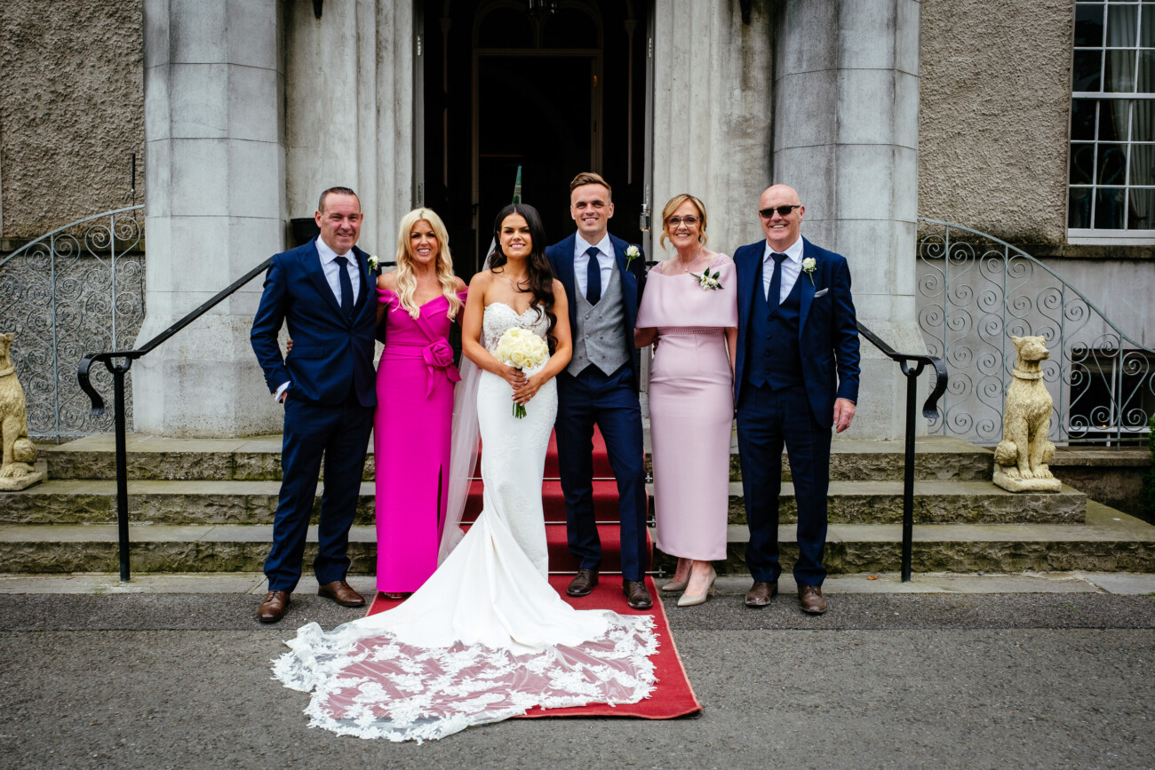 Tara and Darren’s Unforgettable Wedding at Bellingham Castle 131 Six people in formal attire pose together outside a building, with a bride and groom in the centre on a red carpet during their wedding at Bellingham Castle.