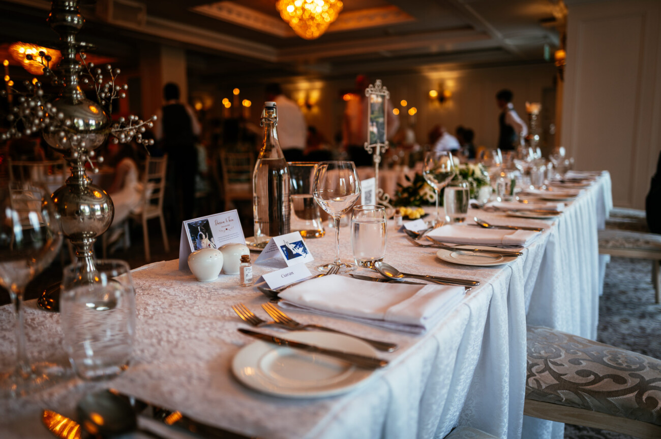 Tara and Darren’s Unforgettable Wedding at Bellingham Castle 132 A formal dining table set with white linen, glassware, plates, cutlery, and name cards in an elegant banqueting hall with chandeliers and blurred guests in the background captures the refined ambience of a wedding at Bellingham Castle.