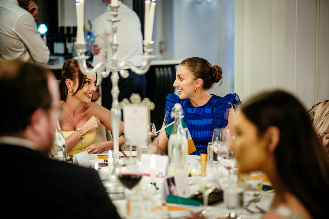 Tara and Darren’s Unforgettable Wedding at Bellingham Castle 133 Two women sit at a formal dinner table, smiling and talking, surrounded by glasses, bottles, candles, and other guests in an elegant indoor setting during a wedding at Bellingham Castle.
