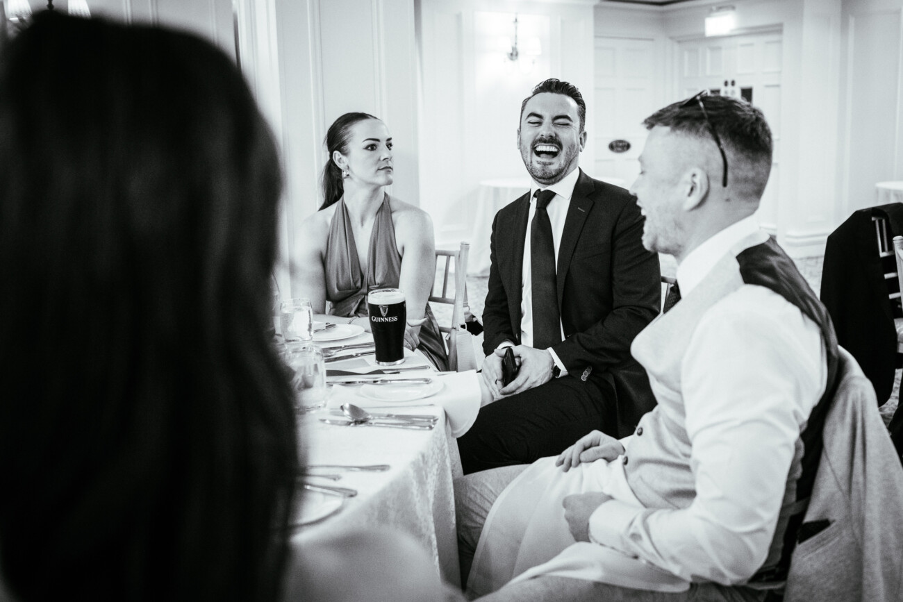 Tara and Darren’s Unforgettable Wedding at Bellingham Castle 134 Four people sit around a table in formal attire; one man is laughing whilst others look on. The setting appears to be a wedding at Bellingham Castle, with the elegant surroundings and warm lighting highlighting the joy of the celebration.