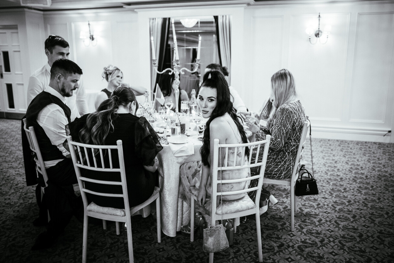 Tara and Darren’s Unforgettable Wedding at Bellingham Castle 135 A group of people sit around a round table set for a meal in an elegant room at a wedding at Bellingham Castle; one woman looks directly at the camera.