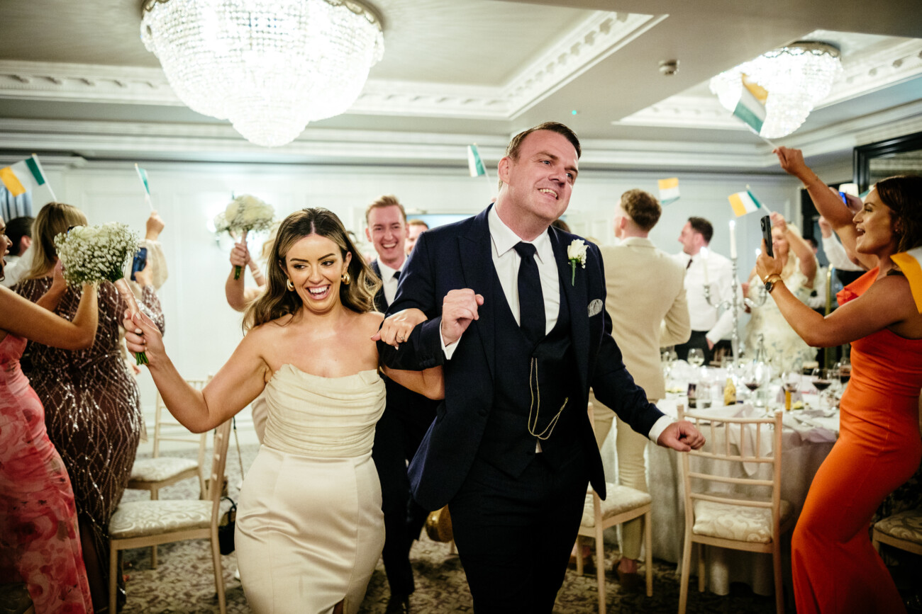 Tara and Darren’s Unforgettable Wedding at Bellingham Castle 136 A bride and groom walk hand in hand through a reception room during their beautiful wedding at Bellingham Castle, as guests wave small flags and celebrate around them.