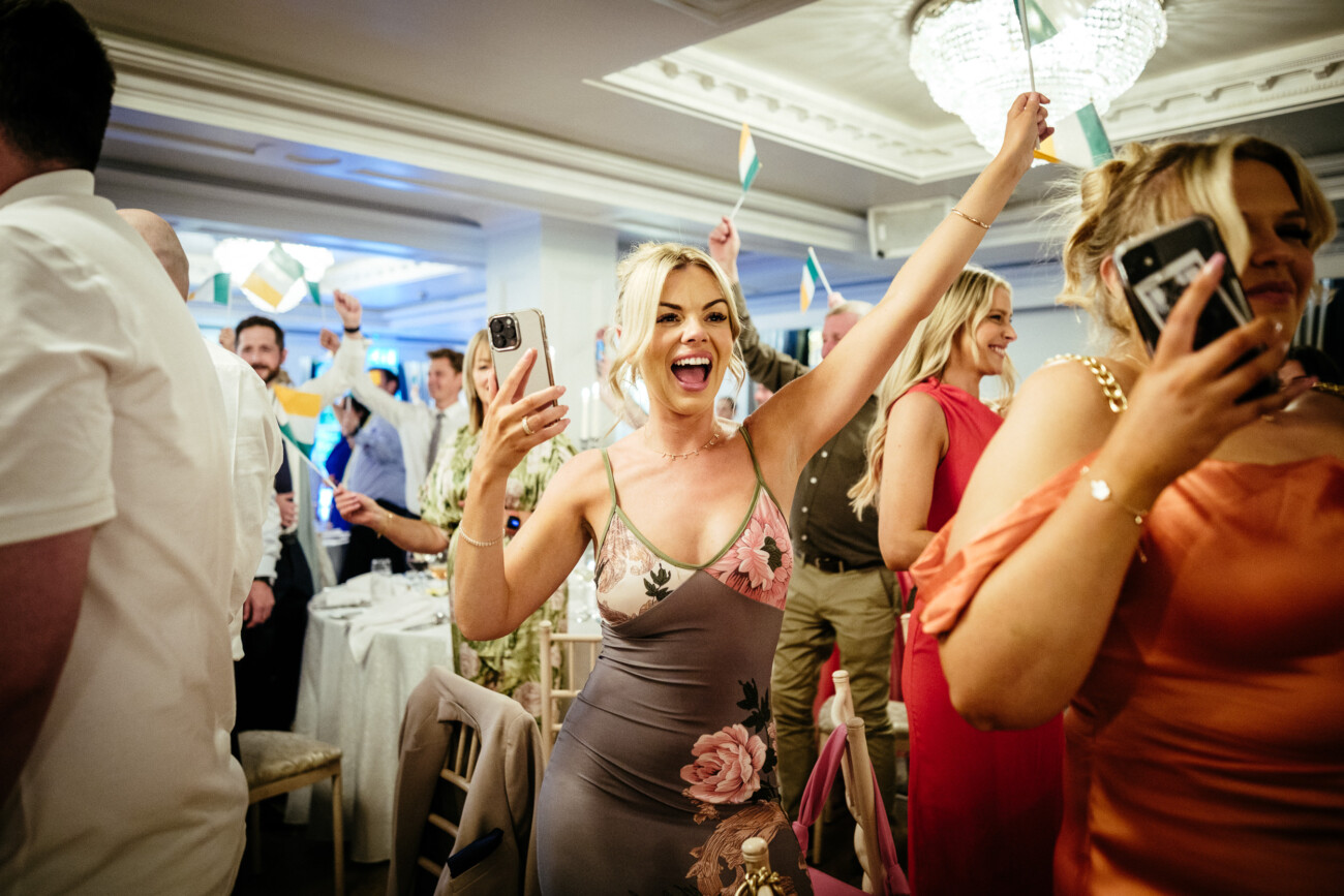 Tara and Darren’s Unforgettable Wedding at Bellingham Castle 137 A woman in a floral dress smiles and raises her arm in a lively, crowded indoor celebration at a wedding at Bellingham Castle, holding a mobile as others wave flags in the background.