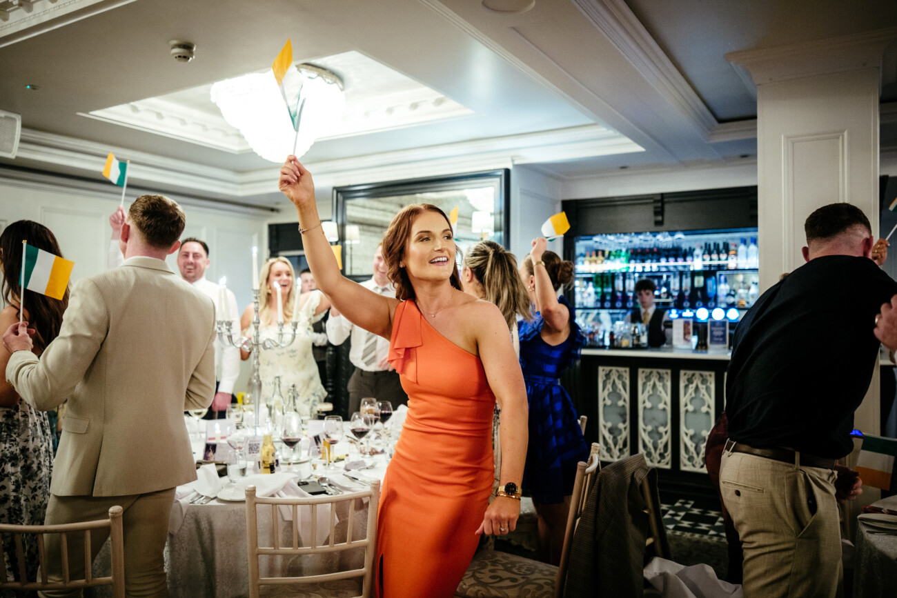 Tara and Darren’s Unforgettable Wedding at Bellingham Castle 138 Woman in an orange dress stands and waves a small Irish flag in a lively, well-lit room during a wedding at Bellingham Castle; other people around her are also holding flags at this elegant formal dining event.