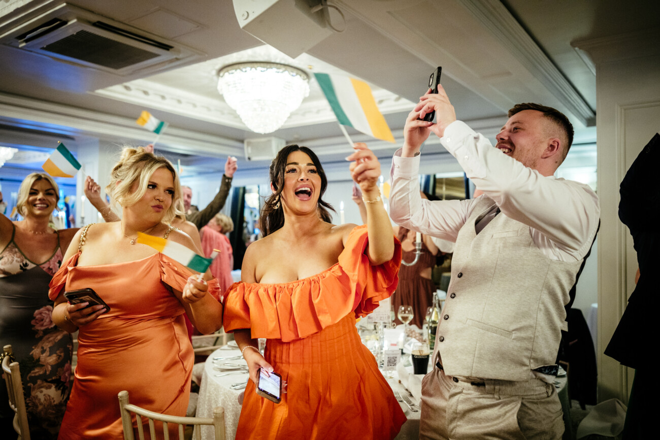 Tara and Darren’s Unforgettable Wedding at Bellingham Castle 139 People in formal attire wave Irish flags and smile at a celebration event, with one person taking a photo on a mobile inside a well-lit banqueting hall during a wedding at Bellingham Castle.