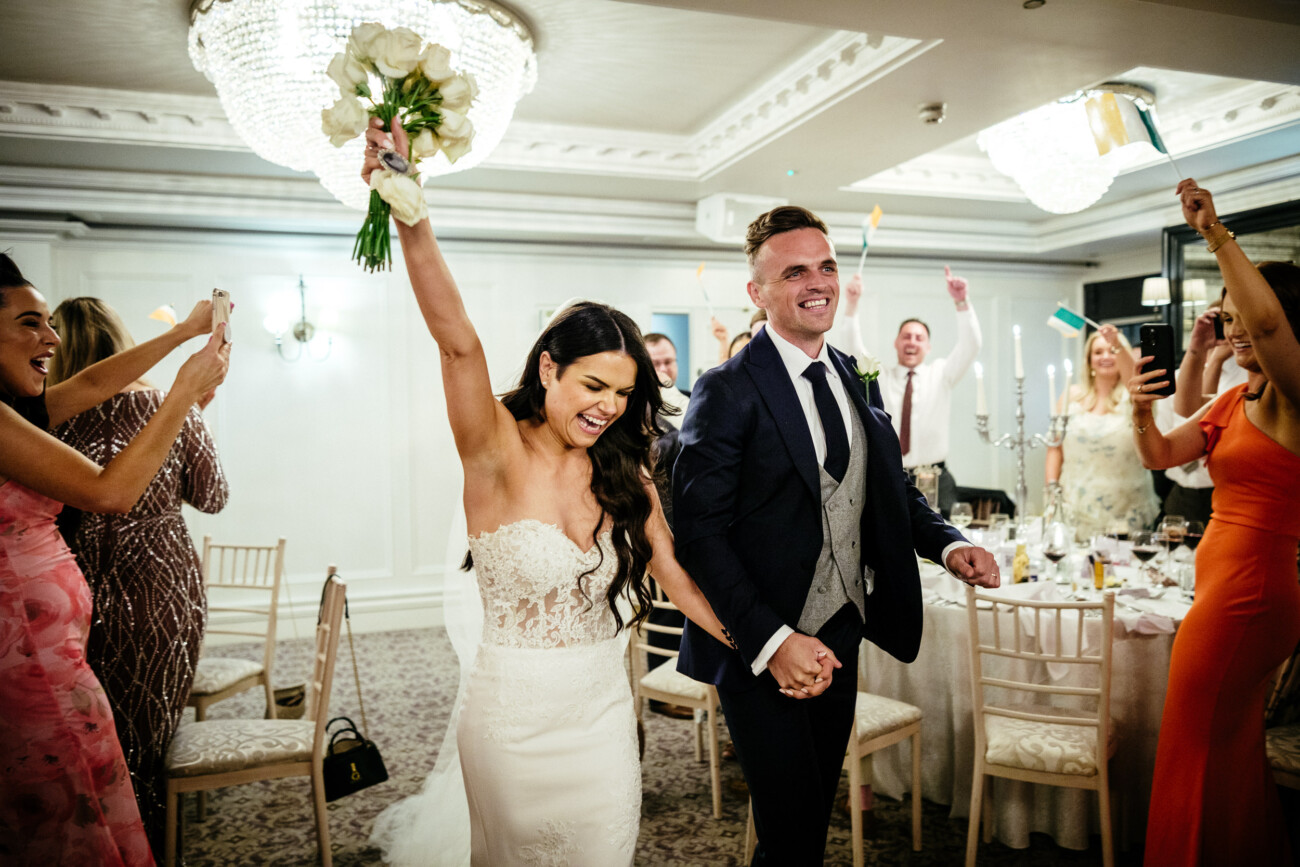 Tara and Darren’s Unforgettable Wedding at Bellingham Castle 140 A bride and groom enter their wedding at Bellingham Castle hand in hand, smiling and celebrating as guests cheer and wave small flags around them.