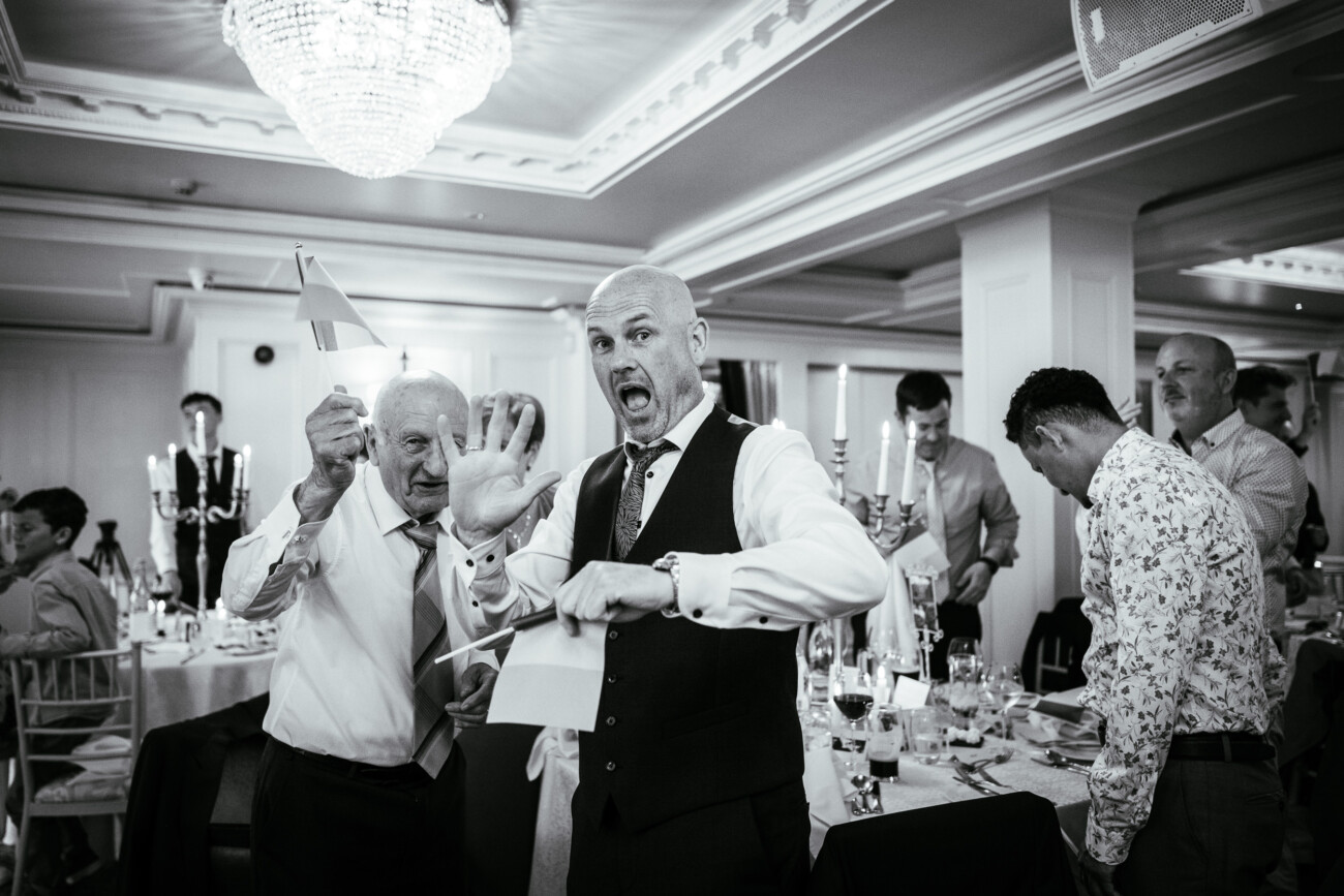 Tara and Darren’s Unforgettable Wedding at Bellingham Castle 141 Two men in formal attire laugh and gesture towards the camera at a lively banquet during a wedding at Bellingham Castle, surrounded by guests seated at decorated tables in a bright, elegant room.