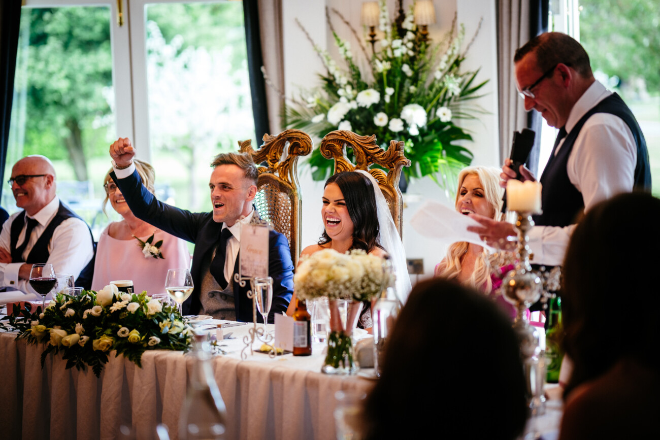 Tara and Darren’s Unforgettable Wedding at Bellingham Castle 142 A wedding at Bellingham Castle comes to life as a bride and groom smile at a beautifully decorated table, surrounded by guests, while a man in glasses gives a heartfelt speech with a microphone.