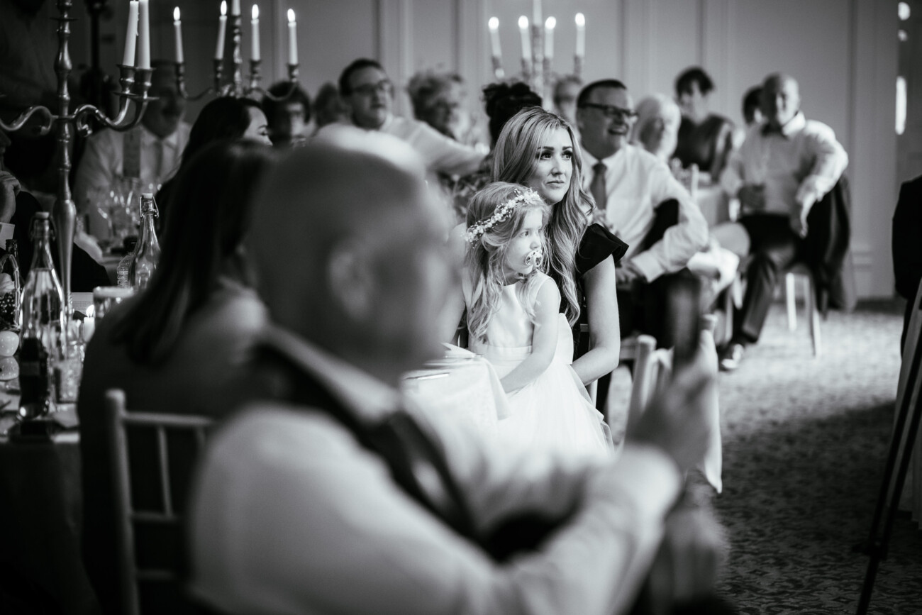 Tara and Darren’s Unforgettable Wedding at Bellingham Castle 143 A group of people sit at tables during a formal event, likely a wedding at Bellingham Castle. In focus, a woman and a young girl in a dress and hairband look towards the front. The atmosphere is calm and attentive.