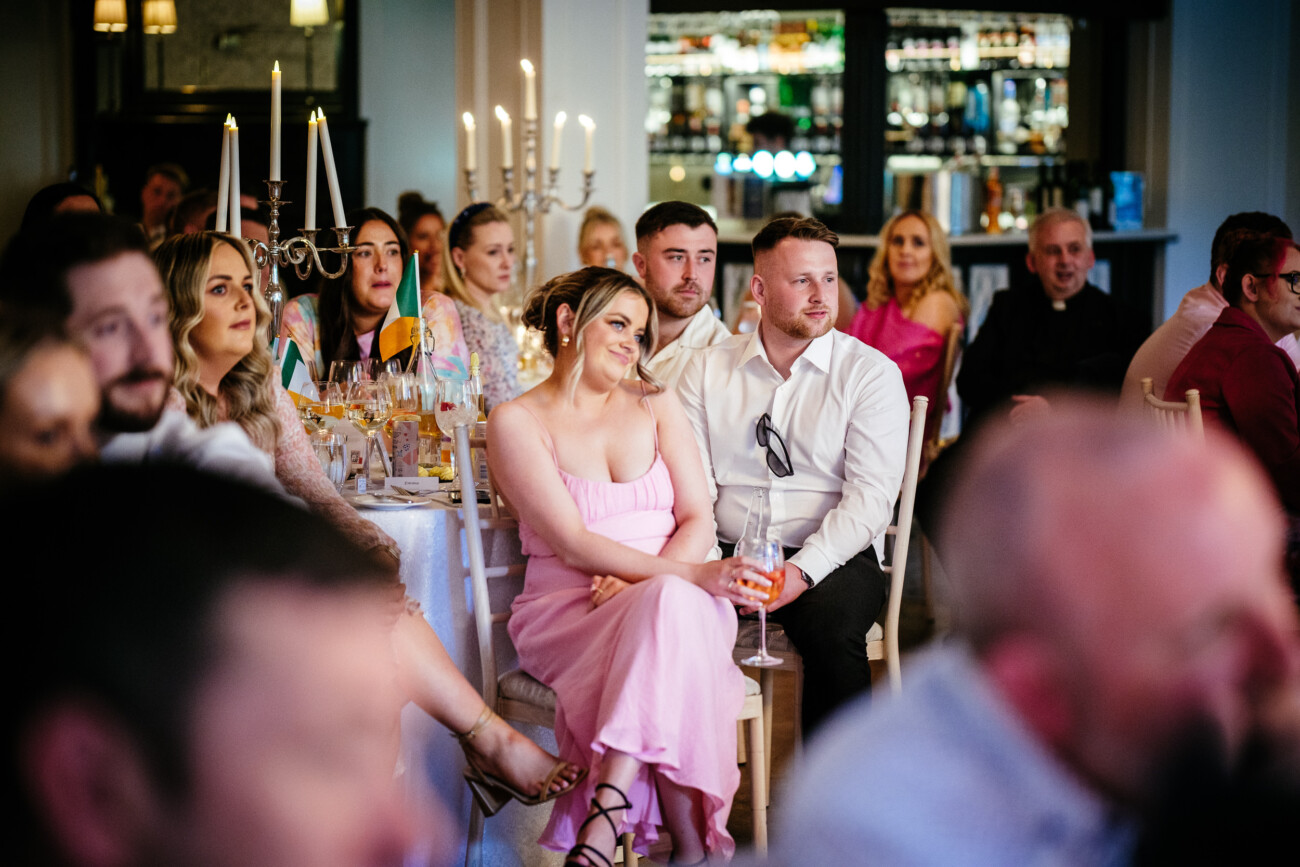Tara and Darren’s Unforgettable Wedding at Bellingham Castle 144 Guests listen attentively during the speeches at the Bellingham Castle wedding reception, with a priest and Irish flag visible in the background.