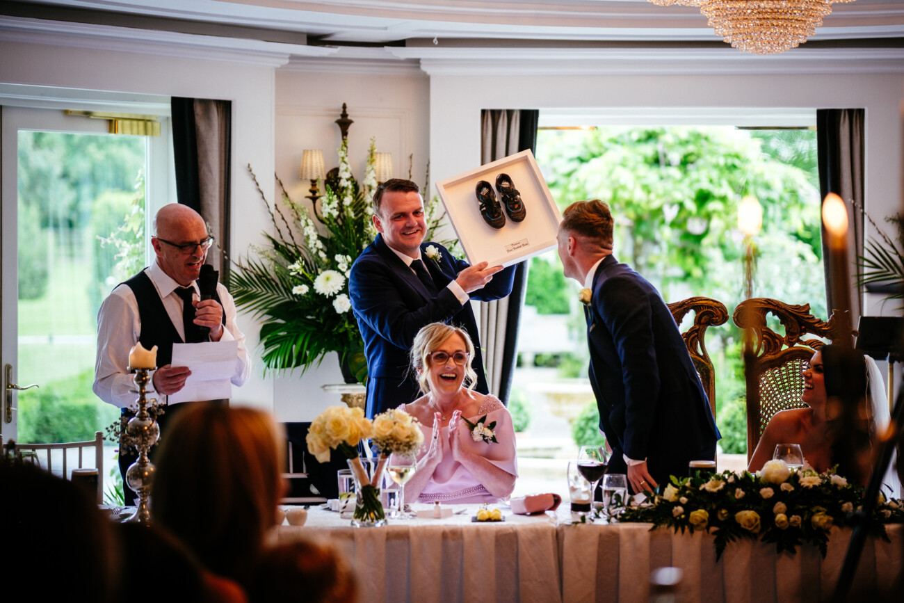 Tara and Darren’s Unforgettable Wedding at Bellingham Castle 145 A man in a suit presents a boxed pair of baby shoes during a formal indoor event at a wedding at Bellingham Castle, whilst guests at a decorated table react with smiles and applause.