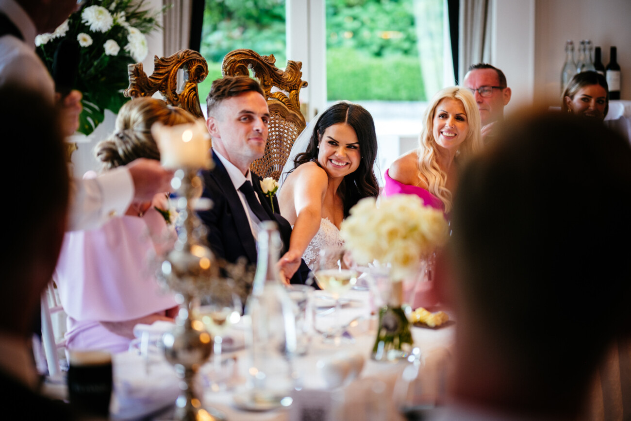 Tara and Darren’s Unforgettable Wedding at Bellingham Castle 146 Bride and groom sit at a decorated table, smiling with guests during their wedding at Bellingham Castle, surrounded by flowers and ornate chairs.