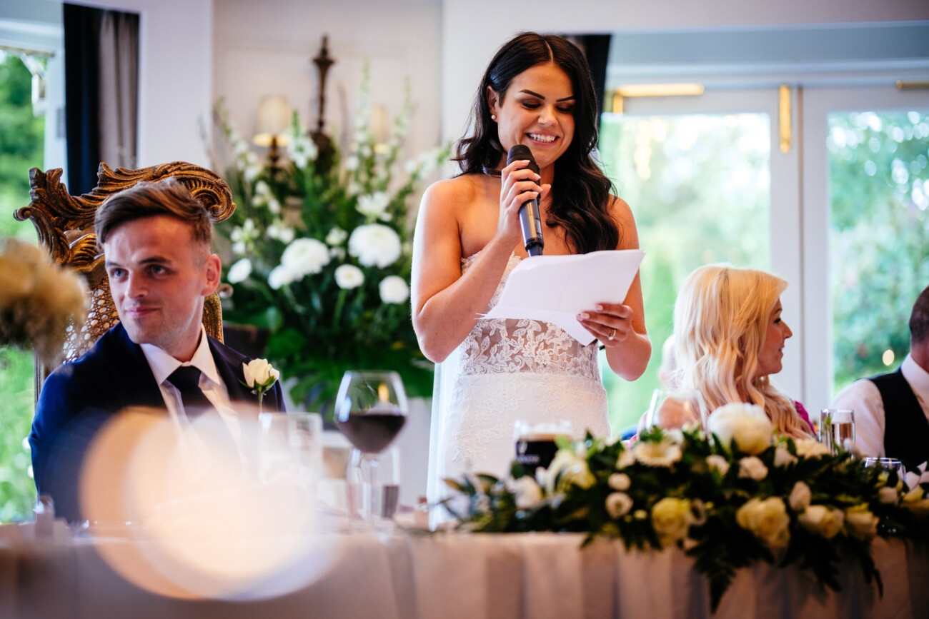 Tara and Darren’s Unforgettable Wedding at Bellingham Castle 147 At a wedding at Bellingham Castle, a woman in a white dress speaks into a microphone whilst holding papers at a decorated table; two people sit beside her.