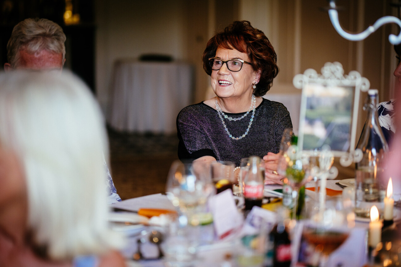 Tara and Darren’s Unforgettable Wedding at Bellingham Castle 148 An older woman wearing glasses and a necklace smiles whilst seated at a beautifully decorated dinner table with food and drinks, enjoying the elegant atmosphere of a wedding at Bellingham Castle.