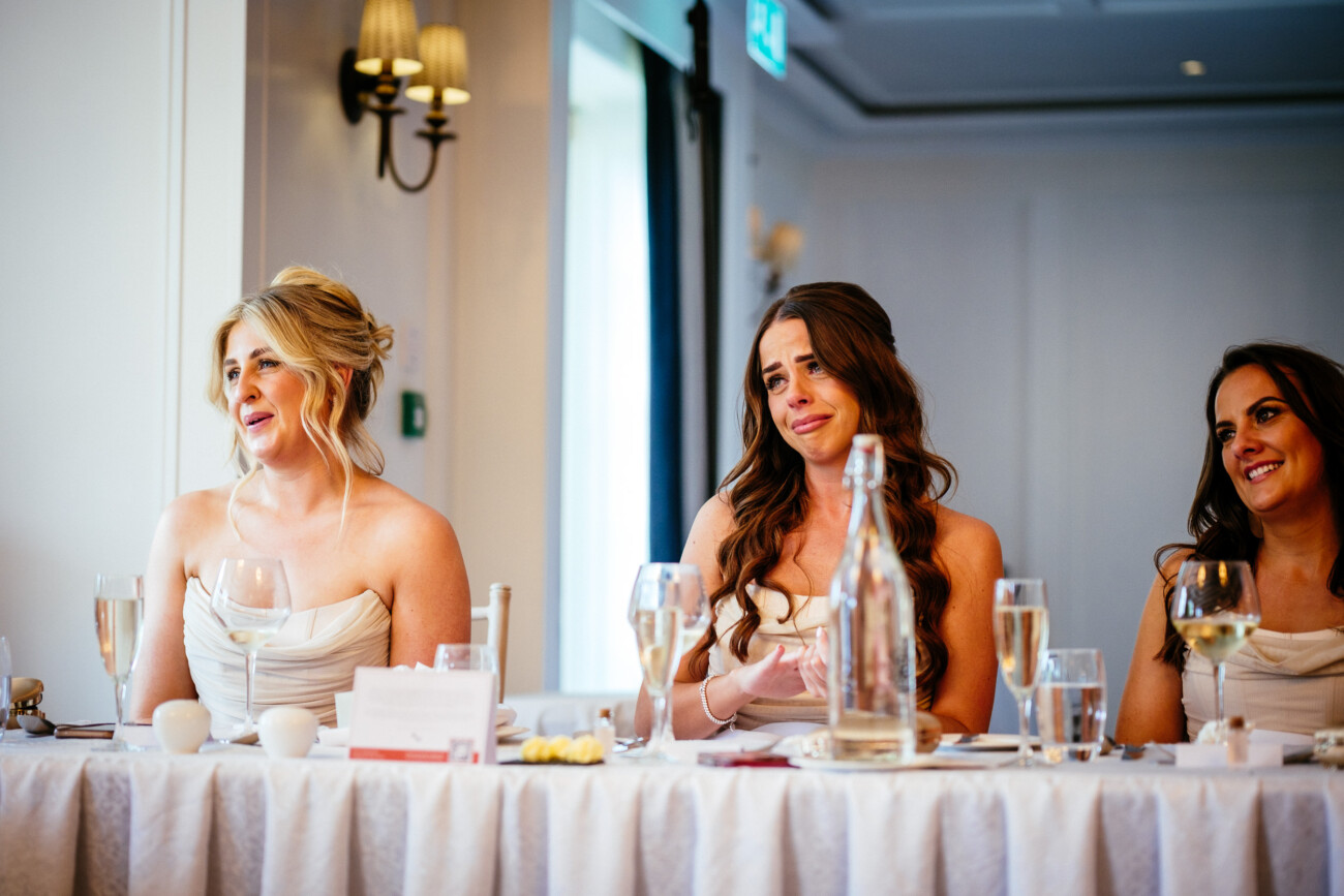 Tara and Darren’s Unforgettable Wedding at Bellingham Castle 149 Three women in light dresses sit at a table with drinks and glassware, displaying emotional reactions—perhaps laughter or tears—during a heartfelt wedding speech or toast at a wedding at Bellingham Castle, in a beautifully well-lit room.